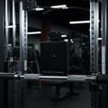 Armaf Club De Nuit Intense Man — Black perfume bottle on a gym barbell, with weight racks in the background.
