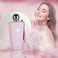 Grandeur Dakota by Grandeur Elite 100ml EDP – Wanita — Botol minyak wangi merah jambu pucat berbentuk faset dengan penutup perak dan juntai, diletakkan di sebelah wanita yang memejamkan mata dan tersenyum, berlatarkan bunga merah jambu dan putih lembut.