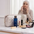 Grandeur Glamour Woman by Grandeur Elite 100ml — A blue perfume bottle with gold accents sits on a marble surface next to a quilted gold makeup bag, lipsticks, and a blush palette, with a woman in a hijab reflected in the background mirror.