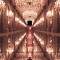 Grandeur Glitz Glam by Grandeur Elite 100 ml for Women — Rose gold glitter perfume bottle with a black cap, reflected on a mirrored surface in a grand hall with chandeliers.