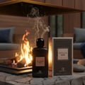 Grandeur Optimum Noir – Botol dan kotak wangian hitam bertekstur dengan aksen emas, diletakkan di atas permukaan batu bersebelahan pendiangan api dengan api dan asap.