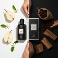 Grandeur Optimum Noir by Grandeur – 100 ml untuk Wanita, botol minyak wangi hitam berlabel putih, dikelilingi hirisan pir, bunga putih, biji kopi dalam guni dan kayu di latar belakang hitam putih terbahagi.