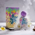 Grandeur Berry Blast by Grandeur Tubbees 50ml Woman — Botol dan kotak pewangi berwarna-warni dengan rekaan cat menitis, dikelilingi bunga putih, batang vanila, dan kaca pecah di atas permukaan marmar, dengan asap nipis.