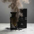 Grandeur Oud Elixir by Grandeur Elite – A cracked glass perfume bottle with dark liquid, surrounded by shattered glass pieces and dark smoke, with the product box in the background.