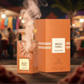 Grandeur Beach Coral by Grandeur 100 ml for Women — A cracked glass perfume bottle with golden accents, emitting smoke, stands next to its orange textured box on a wooden surface, with a blurred market background.
