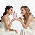 Grandeur Dakota by Grandeur Elite 100ml EDP – Wanita — Dua wanita berbaju satin putih tersenyum sambil memegang botol minyak wangi merah jambu di antara mereka.