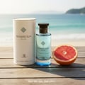 Grandeur Soaked Sun Exclusif by Grandeur Elite EDP 100 ml — A clear glass perfume bottle with blue liquid and a dark cap, next to its white cylindrical box and a halved grapefruit, on a wooden surface with a beach and ocean background.
