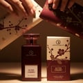 Grandeur Ultra Cherry – A dark red perfume bottle and its box, adorned with cherry blossom and almond motifs, are presented with hands holding decorative wrapping paper.