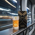Gulf Orchid Sweet Heaven by Gulf Orchid 100ml EDP Unisex — A clear glass perfume bottle with amber liquid and a gold cap, next to its black and gold patterned box, displayed on a metallic railing with blurred city lights in the background.
