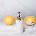 Khadlaj Stellar Musk by Khadlaj 100ml EDP — White bottle with gold accents and pump dispenser, flanked by two yellow fruits on a marble surface with a marble-patterned background.