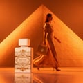 Lattafa Badee Al Oud Honor Glory — Woman in gold dress walks past perfume bottle with pyramid background