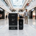 Lattafa Hayaati Men by Lattafa Perfumes 100ml EDP — Black perfume bottle and box displayed on a marble surface in a shopping mall with a man walking in the background.