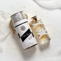 Lattafa Musk Mood by Lattafa 100ml EDP Unisex — Cylindrical white and black patterned box with gold rope handle next to a clear glass perfume bottle with gold cap, both on a white powdery surface with draped white fabric.