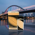 Lattafa Najdia in Gold by Lattafa 100ml Unisex — A perfume bottle and its box are displayed on a reflective surface with a blurred city bridge and skyline in the background.