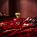 Lattafa Opulent Red by Lattafa 100ml Unisex — Red glass perfume bottle with gold ornate cap, set on red velvet fabric with gold jewelry and rose petals.