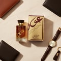 Lattafa Oudain by Lattafa Perfumes 100ml EDP for Men — Amber perfume bottle with gold accents, gold box with maroon text, brown leather wallet, brown leather watch, and keychain on a marble surface.