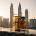 Lattafa Oudain by Lattafa Perfumes 100ml EDP for Men — Gold perfume bottle and box with Arabic calligraphy, set against a blurred cityscape with twin towers at sunset.