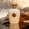 Lattafa Opulent Musk by Lattafa 100ml EDP Unisex — Creamy white perfume bottle with ornate gold cap and sun-like emblem, set against textured brown rocks and a reflective surface.