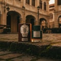 Lattafa Qaaed Intense by Lattafa 100ml EDP — Botol minyak wangi hijau gelap bertekstur dan kotak silindernya yang coklat diletakkan di atas permukaan batu berlumut, dengan bangunan gaya kolonial yang kabur di latar belakang.