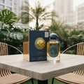 Lattafa Raed Luxe by Lattafa 100ml EDP Unisex — Kotak biru gelap dan emas serta botol minyak wangi kaca jernih bertutup biru dan perincian emas diletakkan di atas meja kayu, dengan tumbuhan hijau subur dan bangunan di latar belakang.