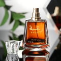 Lattafa Raghba by Lattafa 100ml EDP Unisex — Amber glass perfume bottle with gold Arabic script and 'RAGHBA' text, gold spray nozzle, clear faceted cap beside it, reflected on a dark surface.