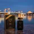 Lattafa Raghba for Men Lattafa 100ml EDP — Black perfume bottle and box with gold accents, displayed on a marble surface with a blurred city bridge and waterfront at dusk in the background.