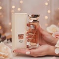 Lattafa Reve Rose VURV By Lattafa 100ml EDP — A woman's hands hold a clear glass perfume bottle with a rose logo and gold cap, next to its cream-colored box, with soft bokeh lights in the background.