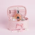 Lattafa Rouat Al Musk by Lattafa 100ml for Women — Pink quilted cosmetic bag open, revealing a clear glass perfume bottle with ornate gold accents and a silver chain, surrounded by pink rose petals and pearls on a pink background.