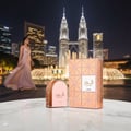 Lattafa Shahd by Lattafa Perfumes 100 ml Women — A woman in a long gown walks past a perfume bottle and box, with the Petronas Twin Towers and fountains in the background at night.