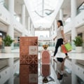Lattafa Shahd by Lattafa Perfumes 100 ml Women — Rose gold patterned box and perfume bottle with gold cap, reflected on a glossy surface in a mall setting.