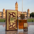 Lattafa Simply Oud by Lattafa 100ml EDP for Men — A gold and maroon patterned box and a clear glass perfume bottle with a wooden cap accent, set on a marble surface with a grand building and clock tower in the background.