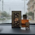 Lattafa Simply Oud 100ml EDP for Men — A glass perfume bottle with amber liquid and a decorative gold and brown cap, next to its matching rectangular box with a geometric design, displayed on a dark surface with raindrops on a window in the background.