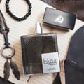 Lattafa Suqraat by Lattafa 100ml EDP Men — Black beaded bracelet, leather necklace with feather charms, and a black perfume cap are arranged around a textured glass perfume bottle on a lined notebook.