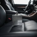 Lattafa Suqraat by Lattafa 100ml EDP Men — A square glass perfume bottle with a textured silver pattern sits on a black leather car seat, with the car's interior visible in the background.