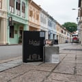 Lattafa Suqraat by Lattafa 100ml EDP Men — Black box and clear glass bottle with silver cap and intricate pattern, displayed on a textured stone pavement with colorful shophouses in the background.