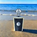 Lattafa Taweel by Lattafa 100ml EDP Unisex — A clear glass perfume bottle with a textured black front panel and a silver cap, placed on a sandy beach with the ocean waves in the background.