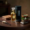 Lattafa Velvet Oud by Lattafa 100ml Unisex — Amber perfume bottle with gold accents, next to its black cylindrical case, a brown leather wallet, a wristwatch, cufflinks, a shaving brush, and a razor on a wooden table.