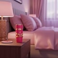 Lattafa Yara Candy by Lattafa 100ml EDP Women — Botol minyak wangi pink dengan perincian emas di atas meja sisi katil kayu bersebelahan lampu dan katil dengan cadar pink.