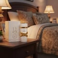Lattafa Yara Moi by Lattafa 100ml EDP Women — White perfume bottle with gold accents and ribbon detail, next to its white embossed box, on a dark wooden nightstand with a bed and lamps in the background.