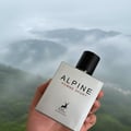 Maison Alhambra Alpine Homme Sport — A hand holds a silver perfume bottle with a black cap against a misty mountain landscape.