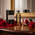 Maison Alhambra B.A.D. Femme by Maison Alhambra 100 ml — A gold and black perfume bottle and its box sit on a wooden surface, surrounded by red velvet fabric and red roses, with a gold-framed mirror in the background.