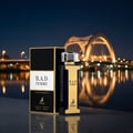 Maison Alhambra B.A.D. Femme by Maison Alhambra 100 ml — A dark blue and gold perfume bottle and its black and gold box are displayed on a reflective surface at night, with a brightly lit bridge in the background.