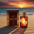 Maison Alhambra Tobacco Your Touch – A golden box and a glass perfume bottle with golden cap sit on a sandy beach at sunset, with the ocean in the background.