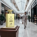 Maison Alhambra Brulant Oud — A tall, rectangular gold perfume bottle with a decorative cap sits on a wooden display stand in a modern shopping mall. A man in a light suit walks in the background.