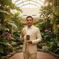 Maison Alhambra Brulant Oud Maison Alhambra for Men 100ml — Man in a cream suit holding a gold perfume bottle and its box in a lush indoor garden setting.