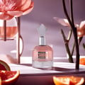 Maison Alhambra Candid by Maison Alhambra 100 ml for Women — Pink perfume bottle with a faceted clear cap, set against a soft purple background with blurred pink flowers and branches. Grapefruit slices are in the foreground.
