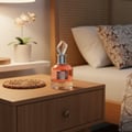 Maison Alhambra Candid by Maison Alhambra 100 ml for Women — Pink perfume bottle with a faceted crystal stopper on a wooden nightstand next to a bed with patterned pillows.