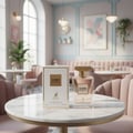 Maison Alhambra Como Moiselle by Maison Alhambra 100ml untuk Wanita — Botol minyak wangi kaca jernih bertutup emas diletakkan di atas meja marmar sebelah kotak berjalur putih emasnya, dalam suasana kafe dengan kerusi baldu merah jambu.