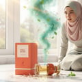 Maison Alhambra Coral Blush 80ml EDP — Botol minyak wangi pecah terbaring, menumpahkan cecair keemasan dan wap hijau pudina berbunga merah jambu, bersebelahan kotak orennya, dengan wanita bertudung memerhati.