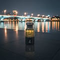 Maison Alhambra Dark Aoud by Maison Alhambra 80ml EDP — Botol minyak wangi kaca gelap dengan penutup bertekstur diletakkan di atas permukaan jubin gelap pada waktu malam, dengan jambatan kabur dan lampu bandar terpantul di air di belakangnya.